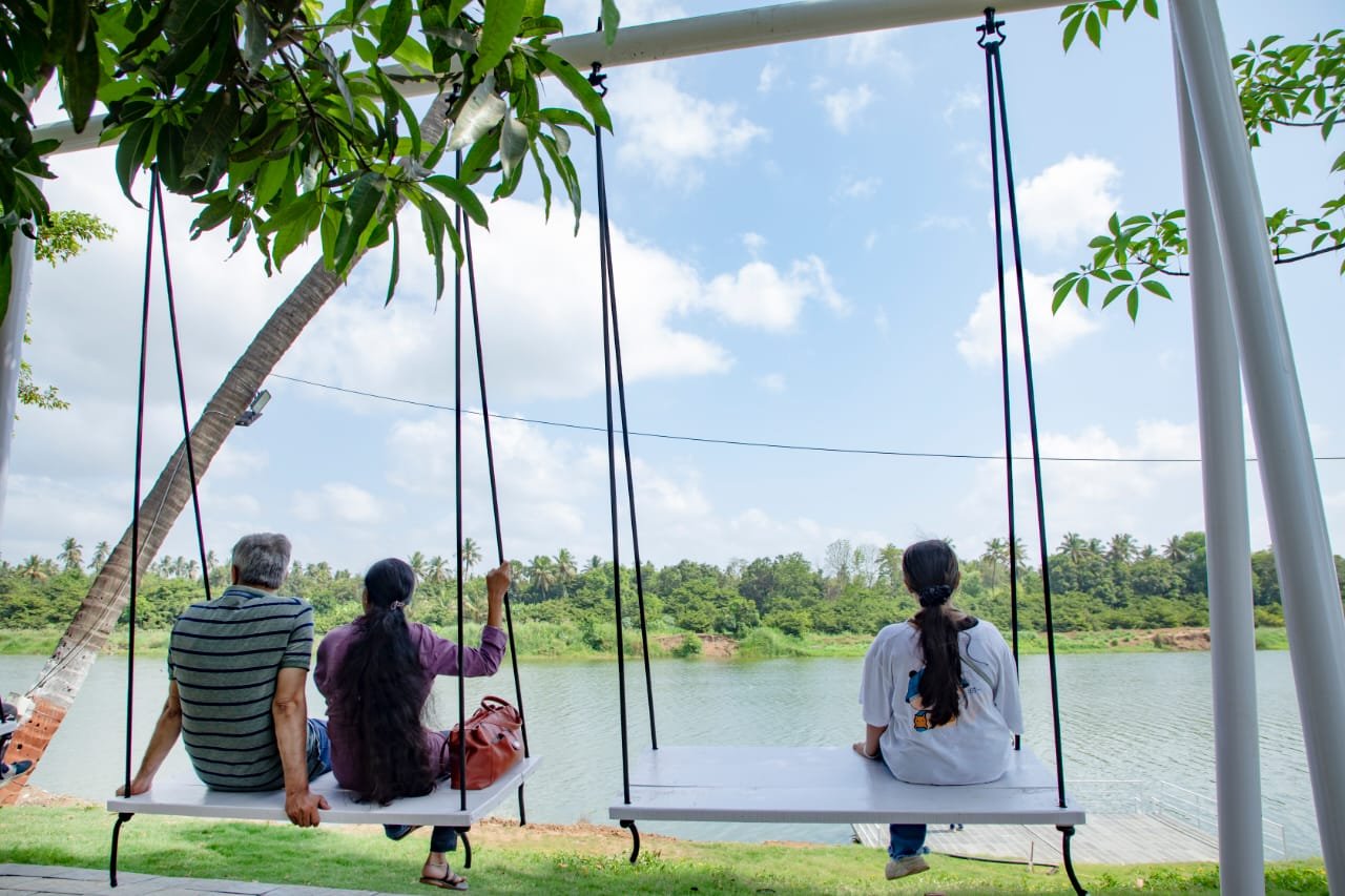 Three people sitting on swings by a serene lake, enjoying a sunny day outdoors